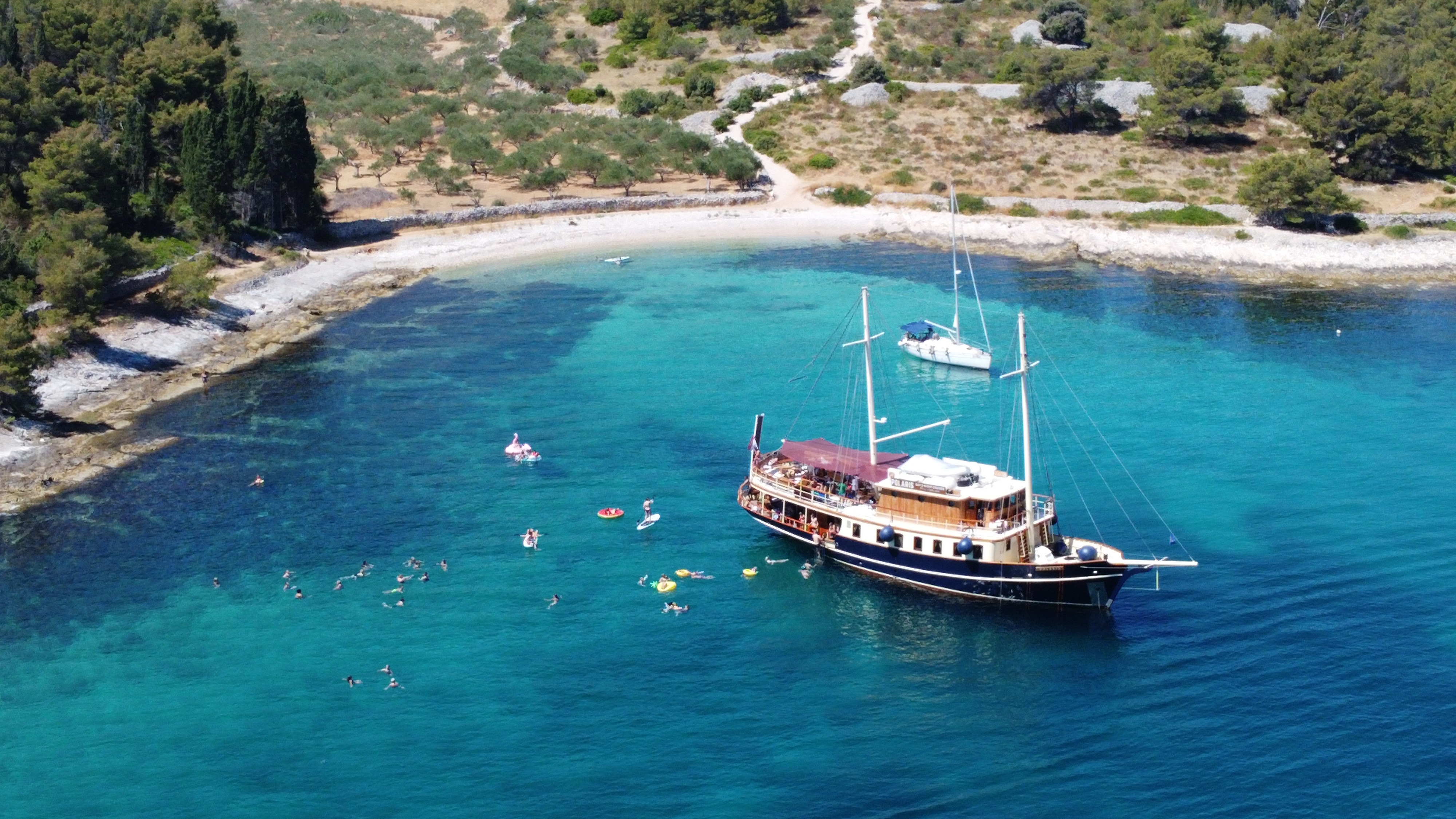 Aerial view of the yacht Polaris anchored in a turquoise Adriatic cove with guests swimming alongside