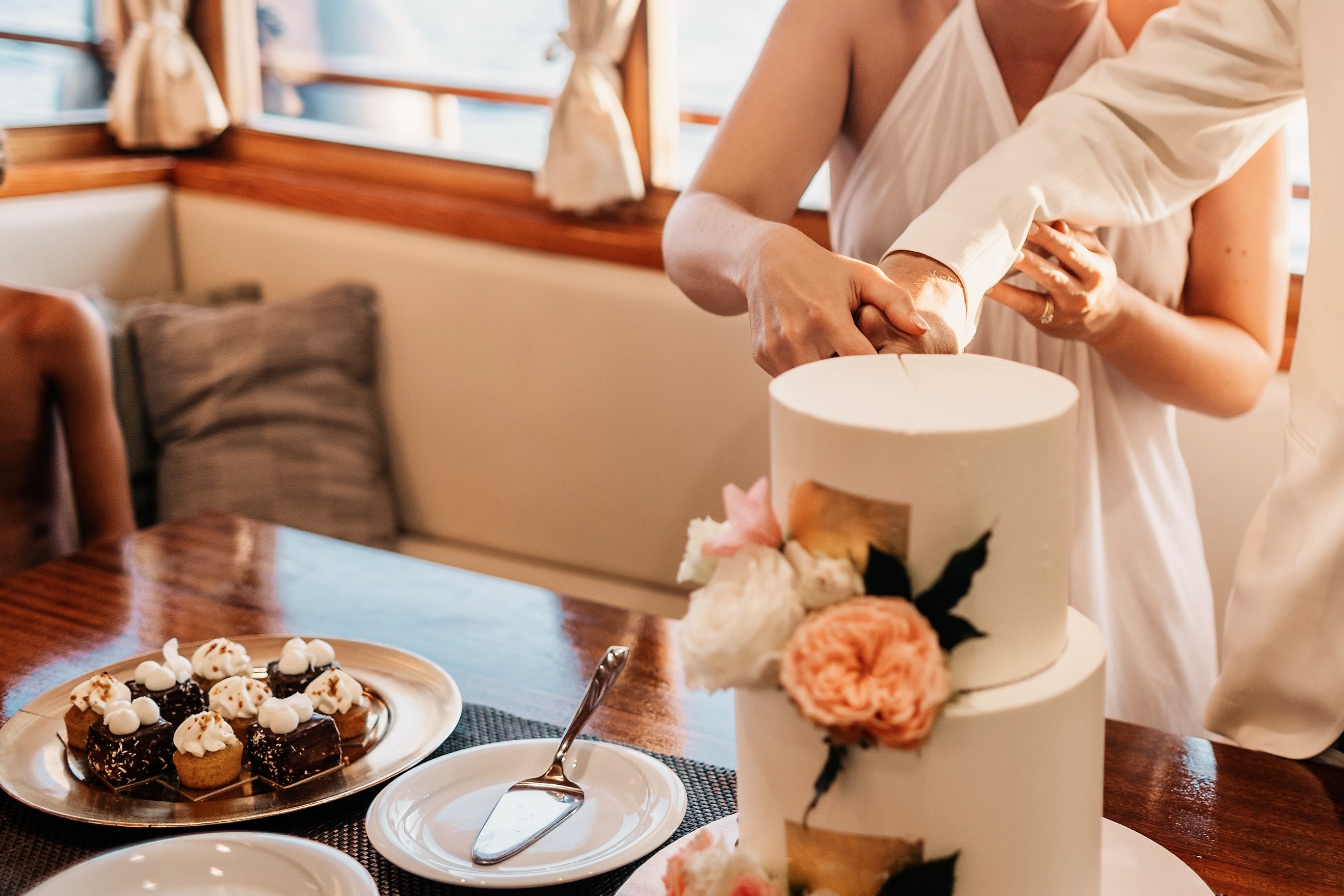 A bride and groom cutting their tiered wedding cake decorated with fresh peonies aboard Polaris, with a platter of petit fours plated beside them
