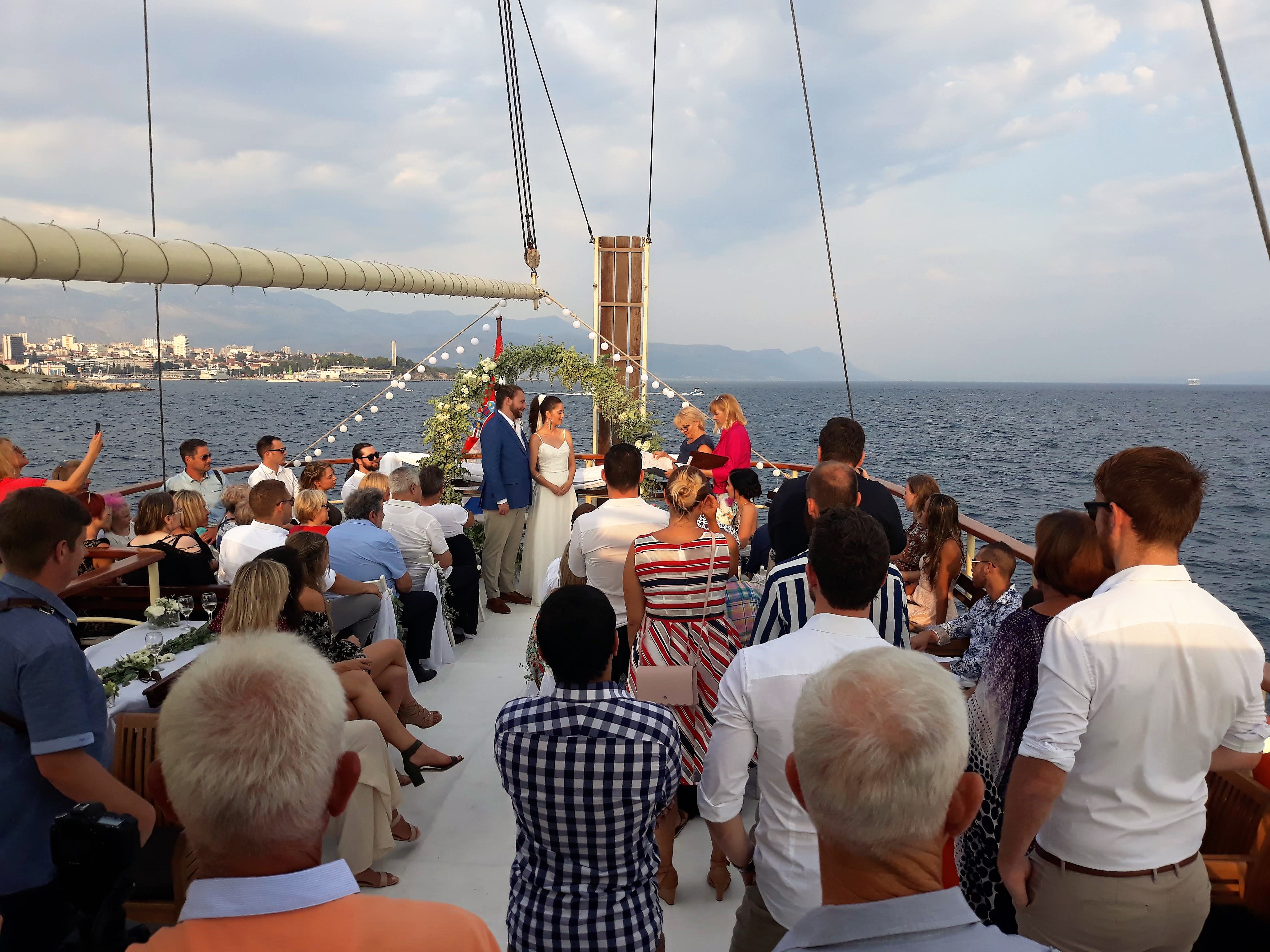 A wedding ceremony on the open deck of Polaris — the bride and groom stand beneath a floral arch strung with lights, surrounded by seated guests, with the Split coastline and mountains behind them