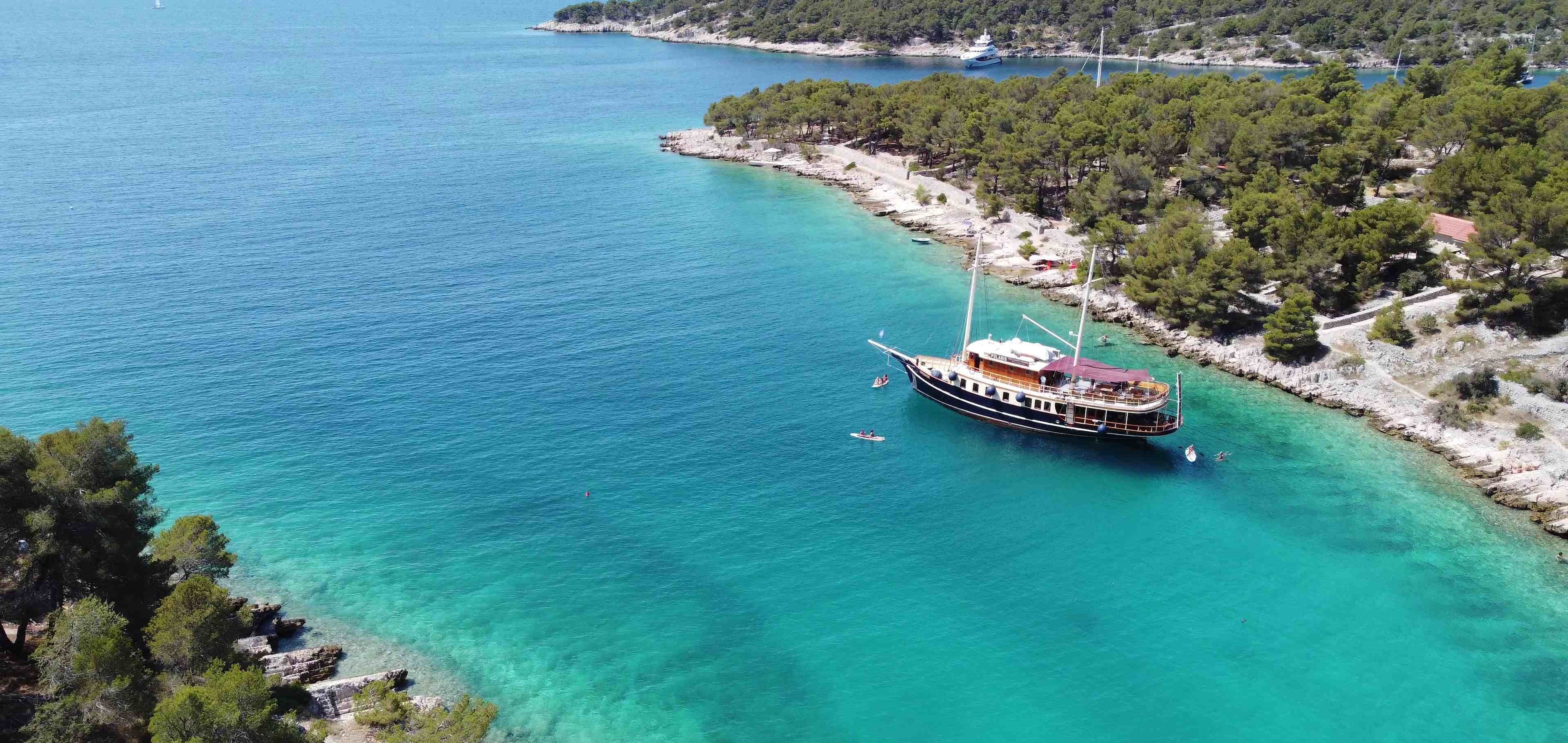 Luftaufnahme der Yacht Polaris vor Anker in einer abgelegenen türkisfarbenen Adriabucht mit Kiefernufern und Paddlern im klaren Wasser daneben