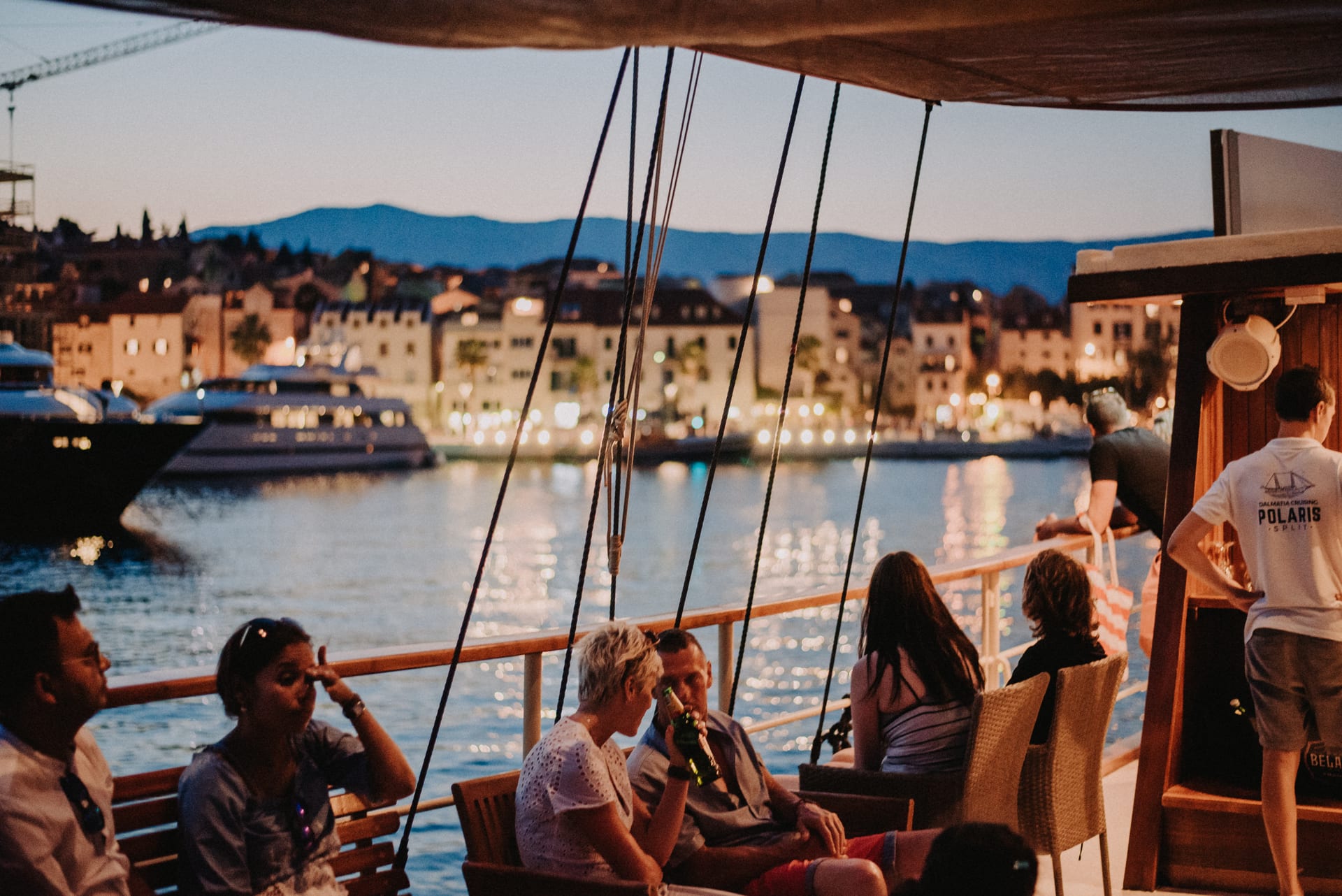 Gäste entspannen in der Dämmerung an Deck mit den Lichtern von Split im Hintergrund