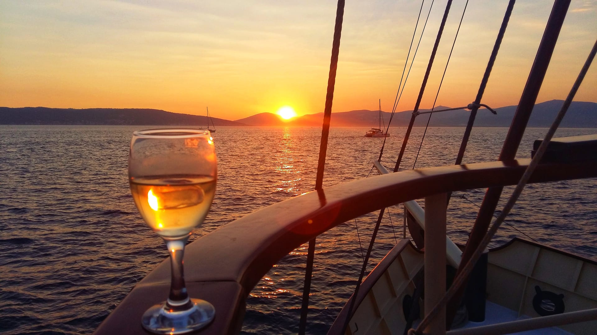 Ein Glas Weißwein auf der Reling, während die Sonne über Inseln und Segelbooten untergeht